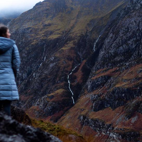 A woman wearing a puffer coat stands overlooking a dramatic, rugged mountain landscape in Glencoe, Scottish Highlands, with thin waterfalls cascading down the slopes. ?? ''
