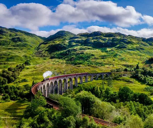 The Jacobite steam train crossing the curved Glenfinnan Viaduct in the Scottish Highlands, surrounded by lush green hills under a blue, cloudy sky. ?? ''